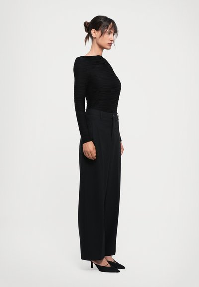 Woman standing sideways wearing a textured black long-sleeve top, wide-leg black pants, and black pointed-toe heels against a plain background.