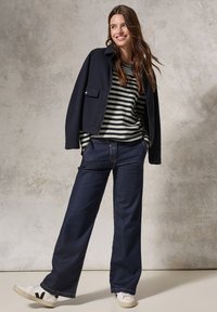 Veste courte marine portée sur une chemise à manches longues rayée noir et blanc, associée à un pantalon en denim sombre ample et des baskets blanches avec des accents noirs.