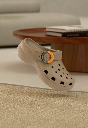 Sabot beige avec des trous de ventilation et une boucle décorative orange et argent flottant au-dessus d'un tapis beige texturé dans un salon moderne.