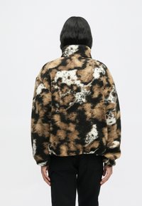 Casaco de fleece em padrão castanho, preto e branco, com gola alta, ombros caídos e um acabamento suave e texturizado.