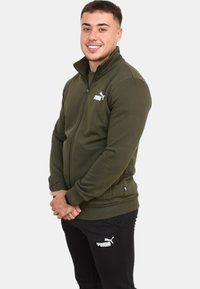 Giacca verde oliva con zip e colletto alto, caratterizzata da un logo Puma bianco sul petto e polsini e orlo a coste. Pantaloni neri con logo.