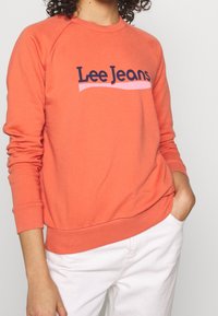 Korálový svetr s žebrovanými rukávy a lemem, se zapracovaným námořnickým logem "Lee Jeans" nad růžovou vlnovkou. Vyrobeno z měkké tkaniny.