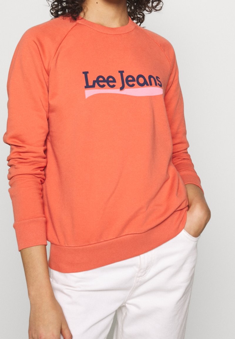 Korálový svetr s žebrovanými rukávy a lemem, se zapracovaným námořnickým logem "Lee Jeans" nad růžovou vlnovkou. Vyrobeno z měkké tkaniny.