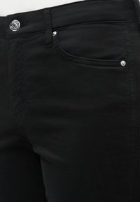 Κοντινή λήψη μαύρων τζιν που δείχνει την εμπρόσθια τσέπη, το θηλάκι της ζώνης και ασημένιο κουμπί και πριτσίνια στο ύφασμα.