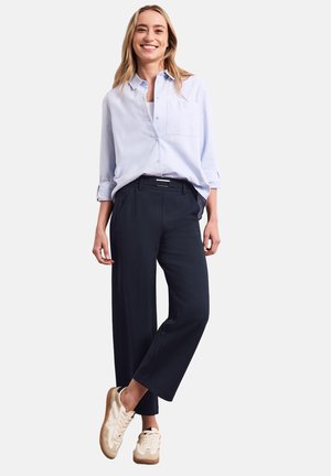 Femme souriante debout, bras détendus, portant une chemise bleu clair à boutons, un pantalon court bleu marine et des baskets beiges sur fond blanc.