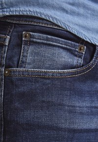 Jeans din denim albastru închis, cu un buzunar din spate vizibil, accentuat de nituri din bronz și cusături mai deschise. Material texturat cu un ușor luciu.
