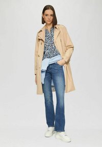 Beige trenchcoat over een blauw bloemenhemde, lichtblauwe gebreide trui om de taille, blauwe jeans en witte chunky sneakers.