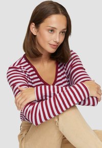Langsleeve gestreept top in bordeauxrood en lichtroze met een diepe V-hals, geribbelde textuur en contrasterende randen. Gecombineerd met beige corduroy broek.