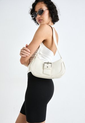 Valge nahast käekott, millel on kõver kuju, reguleeritav rihm ja väike nööbiga detail. Mudel kannab sobivat musta kostüümi.