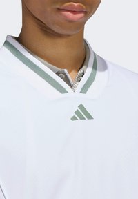 White polo shirt with a green-striped collar, buttoned neck, and a green Adidas logo on the left side. Smooth fabric texture.