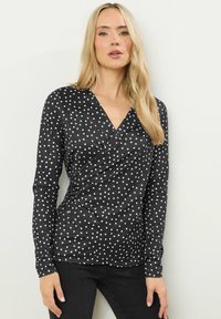 Zwarte lange mouwen wikkelblouse met witte stippen, voorzien van een V-halslijn en een aansluitende pasvorm. Soepele stof met een lichte rek.