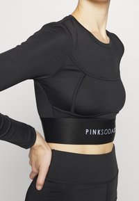 Žena oblečená v černém krátkém topu s dlouhými rukávy a legínách, na elastickém pasu topu je nápis „PINKSODA“.