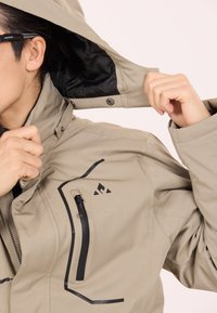 Beige vattentät jacka med hög krage, svarta detaljer, en dragkedjeförsedd bröstficka och en avtagbar huva, med justerbar passform.