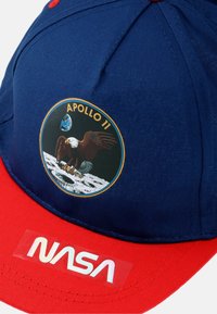 Marineblauwe pet met rode rand, voorzien van een Apollo 11-patch van een adelaar en de Aarde, en een doorzichtige "NASA"-logo aan de voorkant.