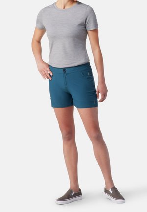 Frau trägt graues kurzärmeliges Shirt, türkisfarbene Shorts mit Knopfverschluss und graue Slipper, steht vor weißem Hintergrund.