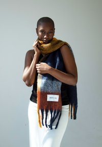 Écharpe en mélange de laine bleue, orange et grise avec des franges. Présente une étiquette blanche portant l'inscription "ARCTIC FOX". Texture douce, forme rectangulaire longue.