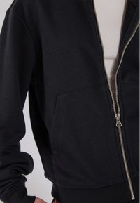 Svart zip-up hettegenser med sølvglidelås, med kengurulomme og ribbede mansjetter. Laget av mykt stoff med en jevn tekstur.