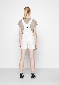 Persoon met blond paardenstaart, beige T-shirt, witte korte tuinbroek met achterzakken en zwarte hoge sneakers, staand tegenover een witte muur.