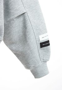 Manga de sudadera gris claro hecha de material suave, con tres etiquetas en blanco y negro. Detalle de puño acanalado en el extremo de la manga.