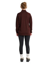 Kvinde med fletbundet hår iført en mørkebrun langærmet top, sorte leggings med "ATW" tekst, hvide sokker og beige vandresko, stående med ryggen til.