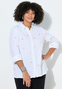 Frau mit lockigem Haar trägt ein weißes Hemd mit Neon-Sternmustern, schwarze Hose, silbernes Armband und Creolen, steht mit der Hand in der Hüfte.