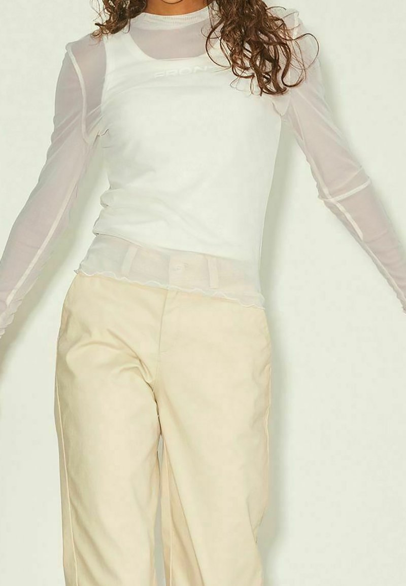 Woman wearing sheer long-sleeve top layered under sleeveless white shirt, paired with high-waisted beige pants against white background.