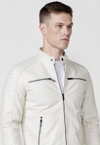 Chaqueta de cuero blanca con cuello alto, bolsillos con cremallera y detalle acolchado en los hombros. Textura suave con acentos de hardware negro.