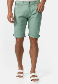 Mintgrüne Baumwollshorts mit umgeschlagenen Säumen, frontalen Taschen und einem Bund mit Knopf und Gürtelschlaufen. Getragen mit weißen Sandalen.