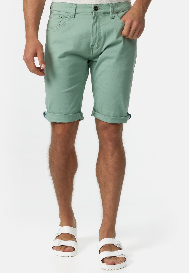 Mintgrüne Baumwollshorts mit umgeschlagenen Säumen, frontalen Taschen und einem Bund mit Knopf und Gürtelschlaufen. Getragen mit weißen Sandalen.