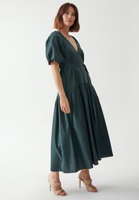 Tume roheline maxi kleit, millel on sügav V-kaelus, puffvarrukad, stimuleeritud talje koos vööga ning kihiline seelik. Kingad on nude tooni.