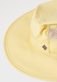 Gul bredbremmet hatt med nettingpanel og stikninger. Kronen er myk, og en logo er vist på siden.