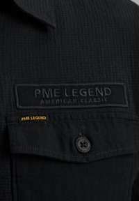 Crna majica s teksturiranim materijalom, "PME LEGEND" logo u podignutom šivanju, i žutim detaljem. Uključuje detalj s crnim dugmetom.