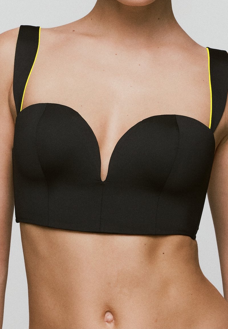 Zwarte cropped top met een diepe V-hals, voorzien van brede bandjes met gele accenten. Glad textuur, gestructureerd ontwerp en moderne silhouette.