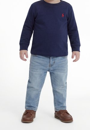 Barn iført navyblå langærmet skjorte med rødt logo, lyseblå jeans og brune læder sko med snørebånd, stående på hvid baggrund.