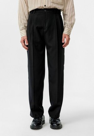 Zwarte op maat gemaakte broek met een recht gesneden ontwerp, geplooide voorkant en riemlussen. Gecombineerd met zwarte schoenen, creëert de gestructureerde stof contrast.