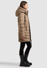 Kurtka puchowa w jasnobrązowym kolorze, z wysokim kołnierzem, pikowaną powierzchnią, wodoodpornym zamkiem błyskawicznym oraz bocznymi kieszeniami. Połączona z czarnymi legginsami i butami.