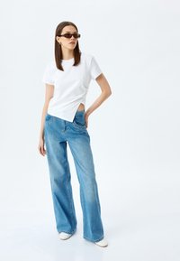 Camiseta blanca de manga corta con un detalle recortado, combinada con unos pantalones vaqueros de pierna ancha en azul claro y complementada con unas gafas de sol rectangulares marrones.