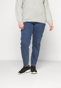 Suéter de malha cinza-claro com mangas longas, combinado com jeans de ganga azul-escuro e sapatilhas pretas com solas brancas.