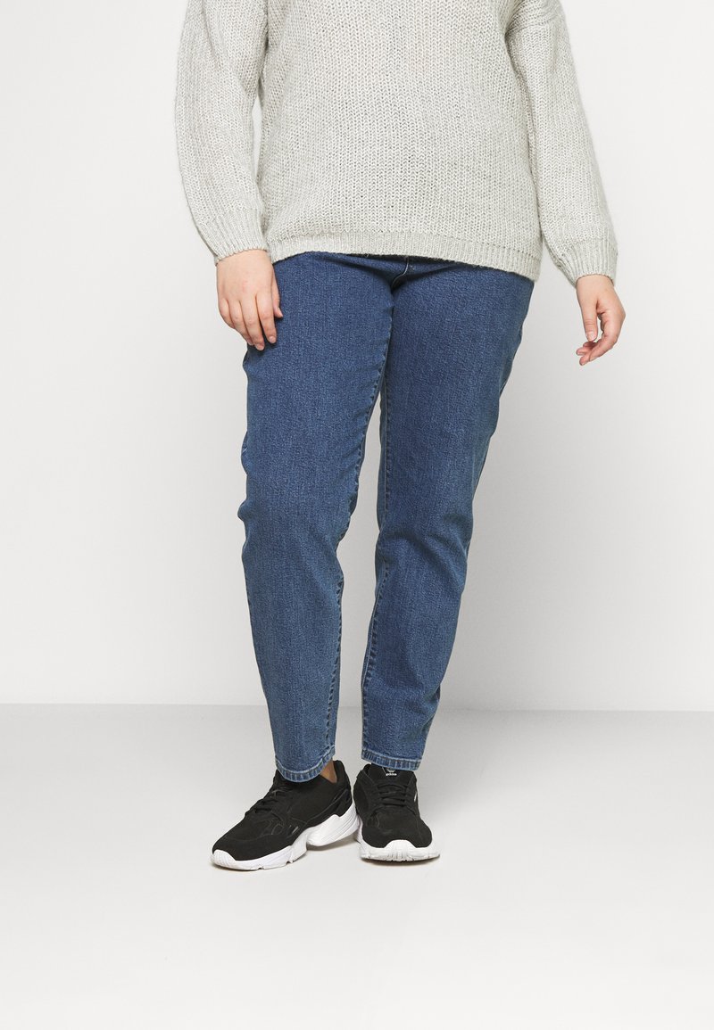 Suéter de malha cinza-claro com mangas longas, combinado com jeans de ganga azul-escuro e sapatilhas pretas com solas brancas.