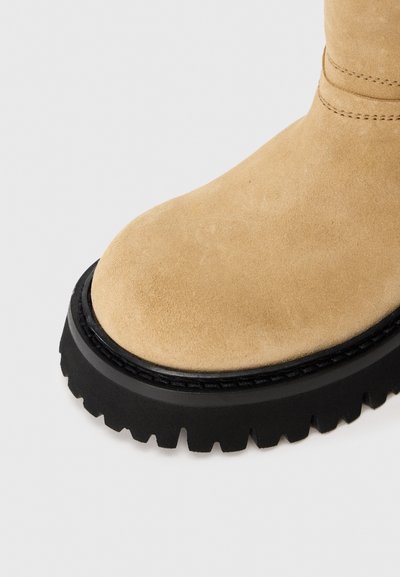 Beżowy zamszowy czubek buta z czarną, masywną gumową podeszwą oraz szczegółowym przeszyciem na białym tle.