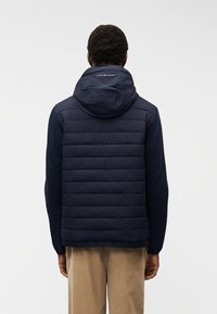 Άνδρας που φοράει σκούρο μπλε επενδεδυμένο μπουφάν με κουκούλα, στέκεται με την πλάτη, συνδυασμένο με μπεζ παντελόνι, μπροστά σε απλό φωτεινό φόντο.