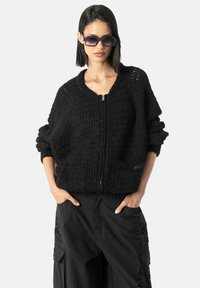 Pulover negru tricotat, cu fermoar, având un model texturat, mâneci largi și decolteu în V. Purtați peste pantaloni cargo negri; modelul poartă, de asemenea, ochei de soare închiși la culoare.