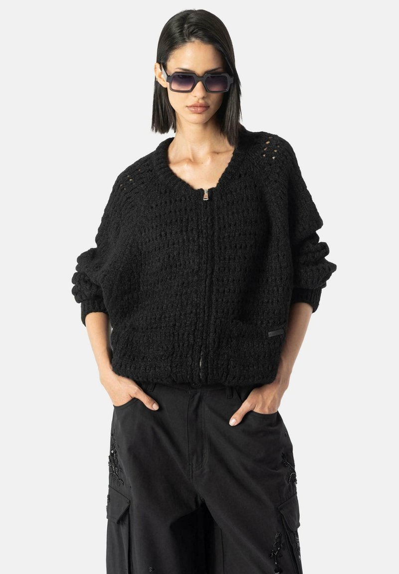 Pulover negru tricotat, cu fermoar, având un model texturat, mâneci largi și decolteu în V. Purtați peste pantaloni cargo negri; modelul poartă, de asemenea, ochei de soare închiși la culoare.