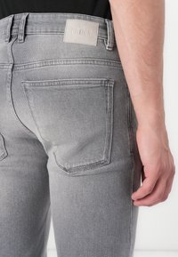 Grå denim jeans med en nærbildevisning av baksiden. Funksjoner inkluderer et lærmerke med "ORYKORN," og to baklommer. Jevnt tekstur.