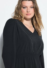 Donna con capelli biondi ondulati indossa un vestito nero con maniche a sbuffo e decorazioni a V con borchie su uno sfondo bianco.