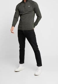 Ciemnozielony, długi sweter z półzamkowym kołnierzem, wykonany z miękkiego materiału, zestawiony z czarnymi, dopasowanymi jeansami i białymi trampkami.