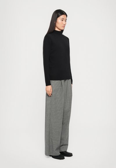 Schwarzer Longsleeve-Turtleneck-Pullover kombiniert mit locker sitzenden grauen Hahnentritthosen und schwarzen Stiefeletten. Glattes Material mit weicher Textur.