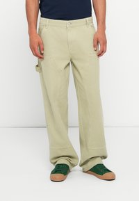 Pantalons cargo larges beige clair avec poches avant et poche latérale, fabriqués en tissu durable ; associés à des baskets vertes et un haut bleu marine foncé.