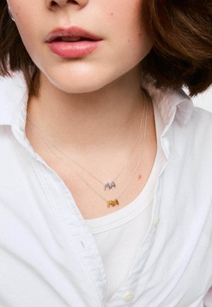Gros plan d'une femme portant deux colliers superposés avec des pendentifs lettres "M" et "A" en argent et or, col de chemise blanc visible.