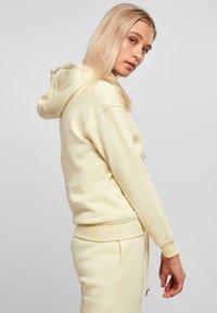 Lys gul hettegenser laget av mykt stoff med snorhette og ribbestrikkede ermer. Designet har en avslappet passform og minimal detaljering.